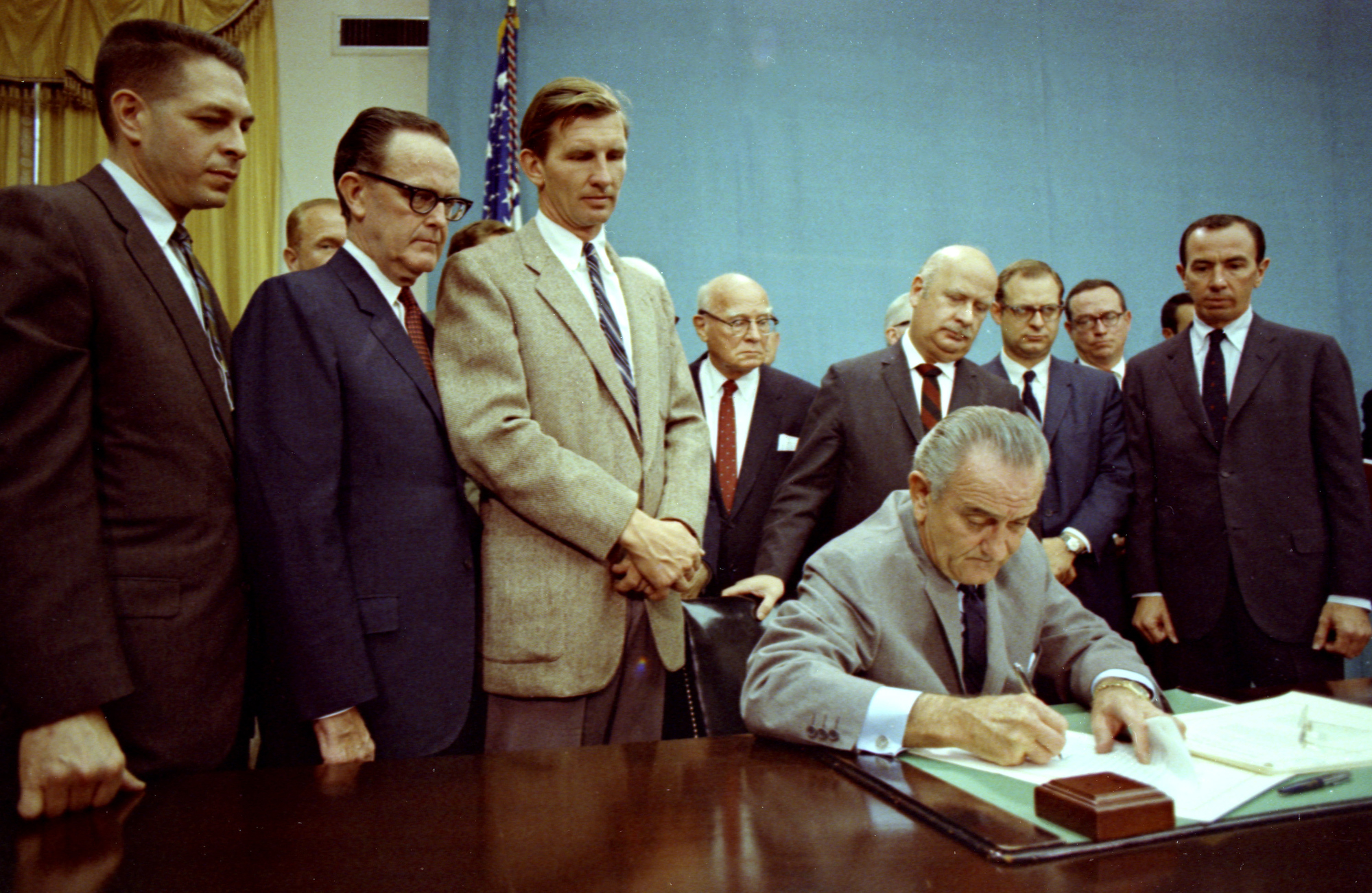 Lyndon Johnson signs a document at a table while a group of officials stands behind him.