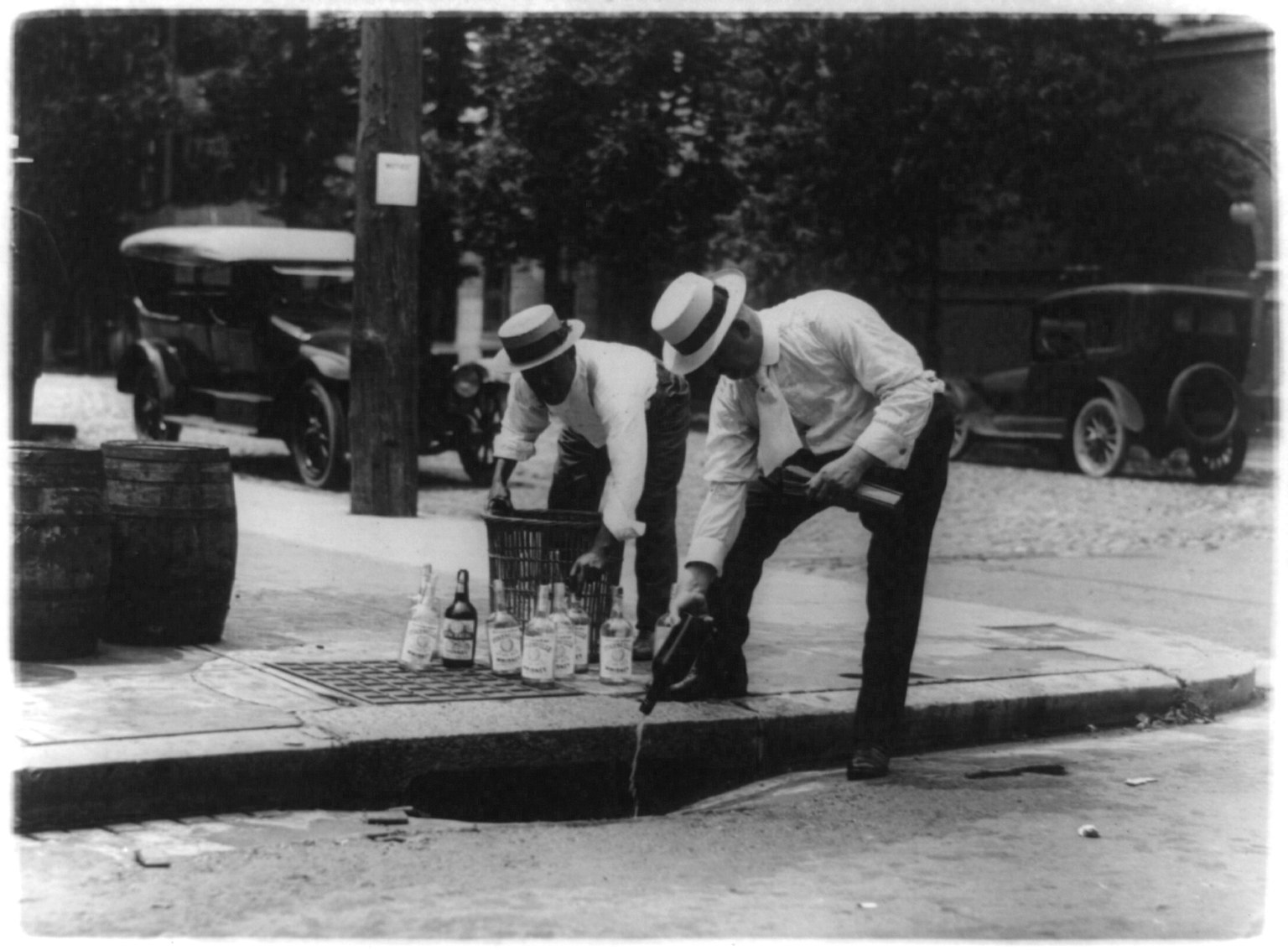 Two men in straw hats empty bottles into a street drain, with barrels and parked cars behind them.