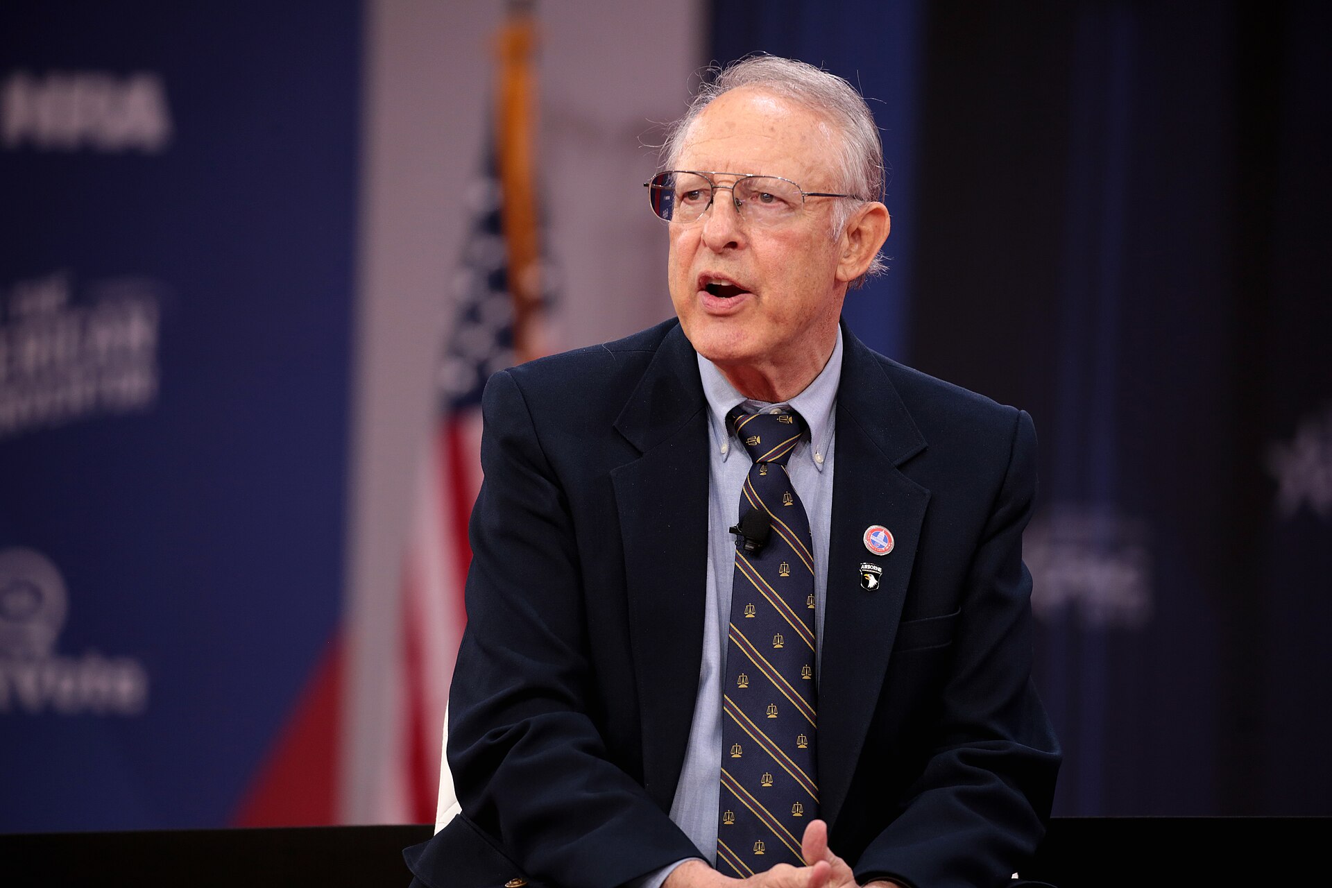 Dick Heller, wearing glasses and a patterned tie, speaks while seated in front of a blue backdrop.