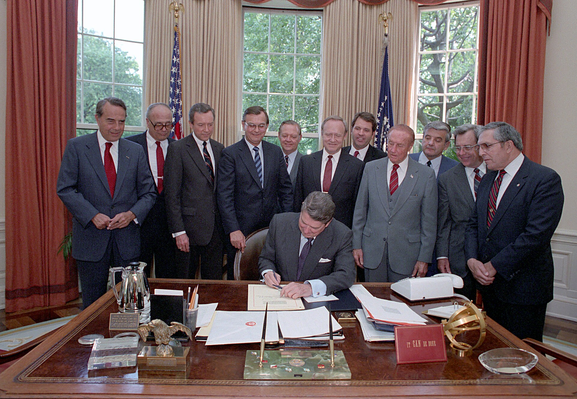 Ronald Reagan signs a document at his Oval Office desk as members of Congress stand behind him.