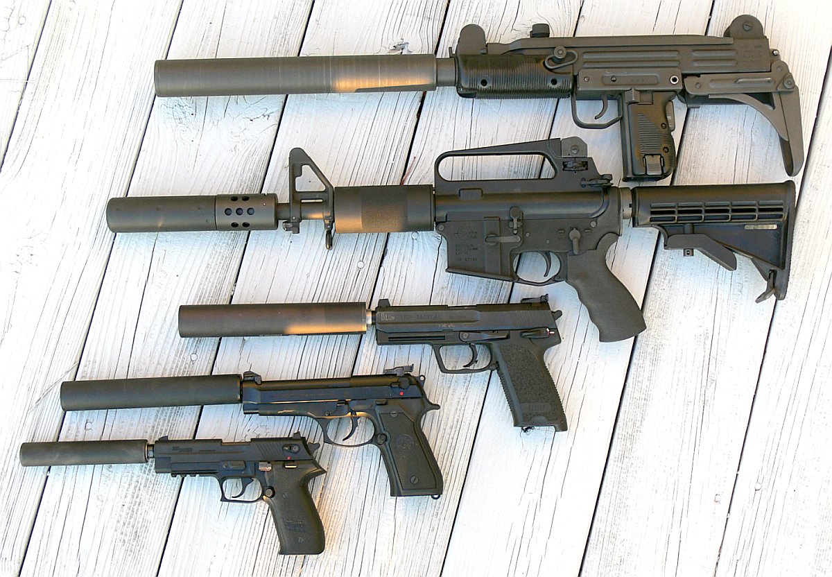 Five firearms with suppressors attached are arranged on wooden boards.