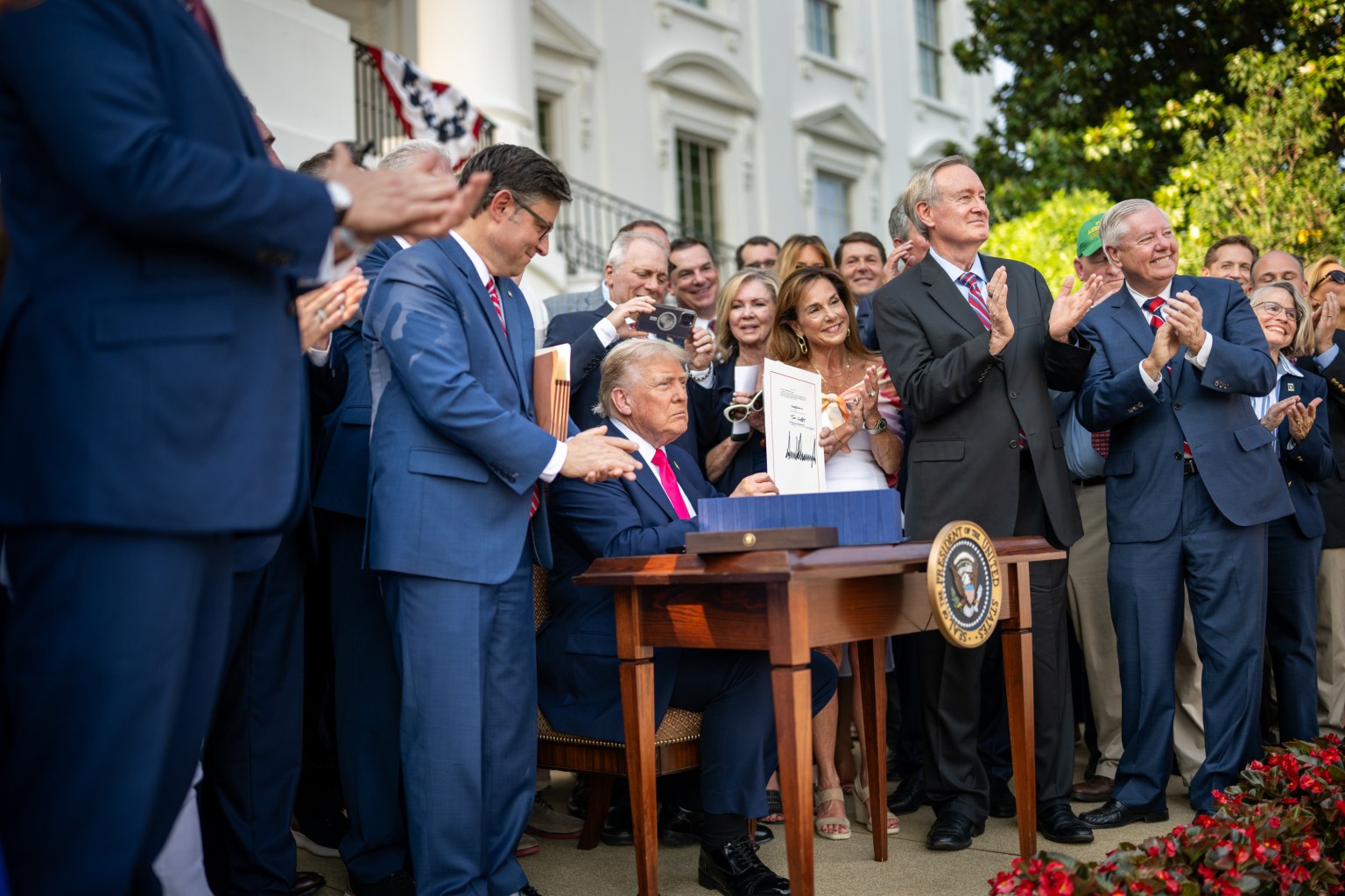 Donald Trump holds up the signed bill at an outdoor desk as officials standing behind him applaud.